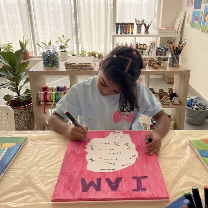 Young girl writing on a pink 'I AM' canvas in a bright art studio.