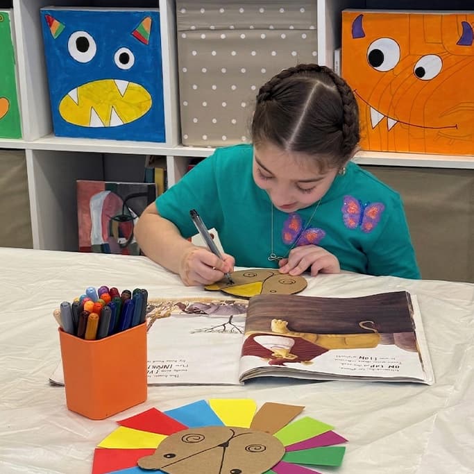 Young girl with braided hair draws on a cardboard lion craft beside an open book.